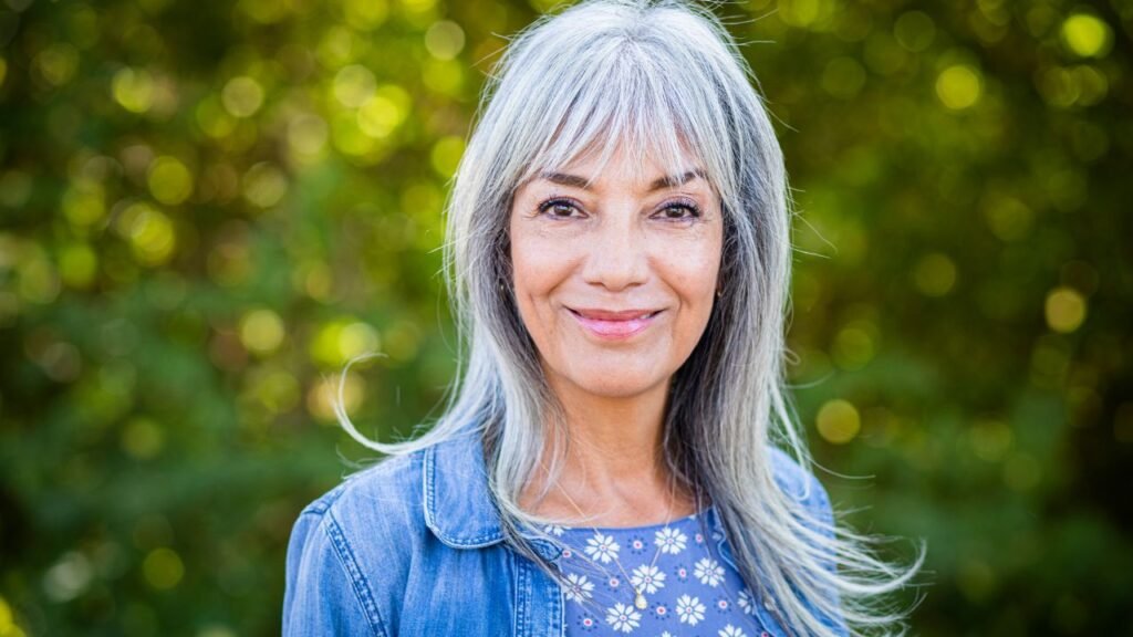 Portrait d'une femme souriante aux cheveux blancs avec un fond de verdure flou.